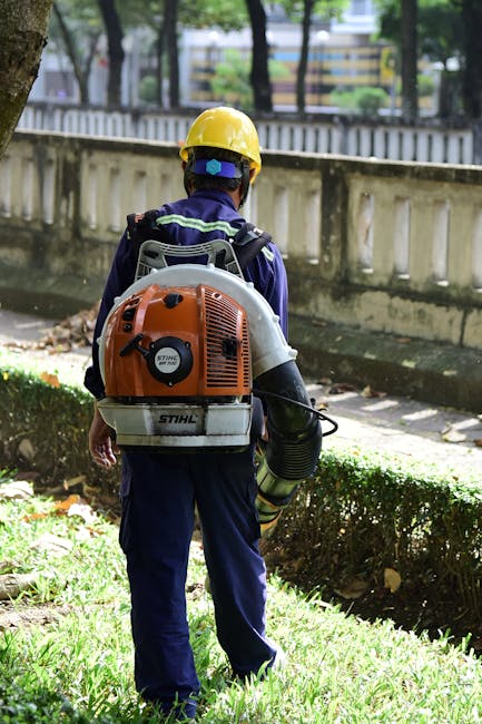 A person wearing a dark long-sleeve shirt and a black wide-brimmed hat is seen from behind, trimming a dense, lush green hedge with a pair of long-handled hedge shears. The hedge is composed of small,
